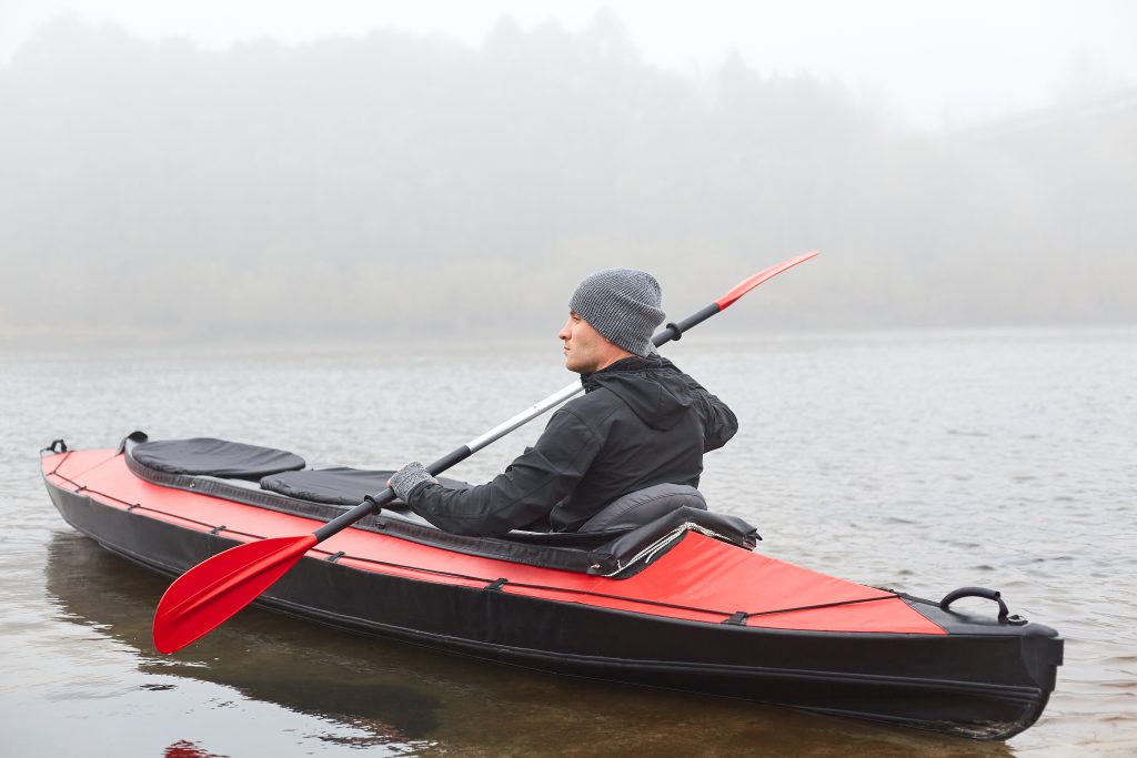Side view of man kayaking on river in cold autumn cloudy day, kayaking, sports and recreation, male wearing black jacket rowing boat, water sport, leisure time in open fresh air.